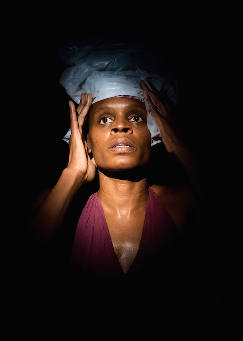 Okwui holds a piece of plastic around her hairline like a scarf. Her skin glistens with sweat.